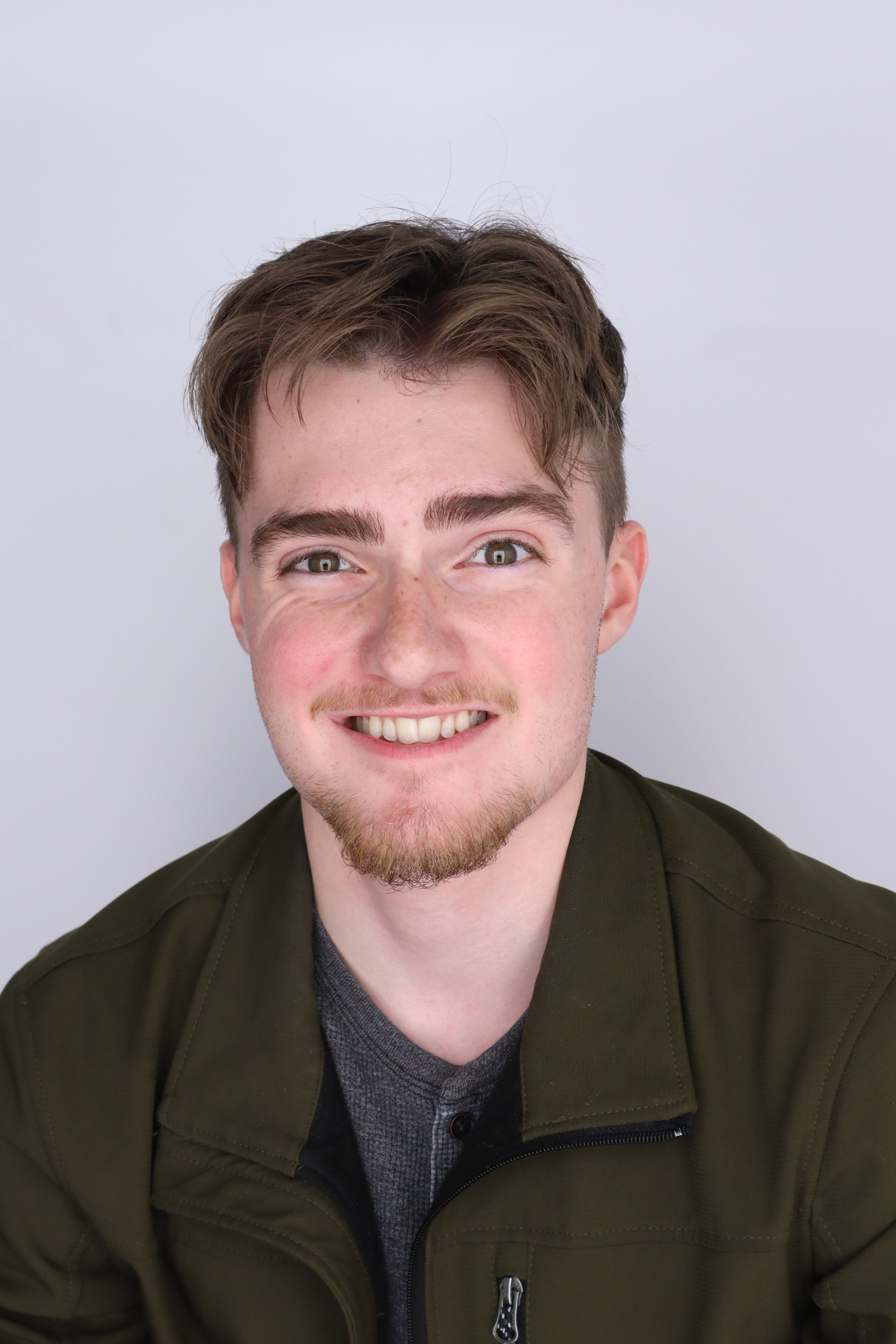 Mitchell Collins smiles, he is sitting in front of a white background and is wearing a dark green jacket.