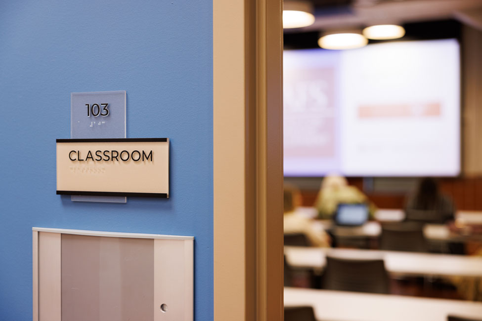 Exterior of entry to classroom. Classroom wall sign is in the foreground with CED students sitting in a classroom in background.