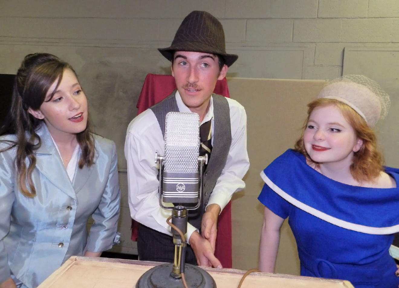 Three singers in 1940s dress surround a large radio microphone