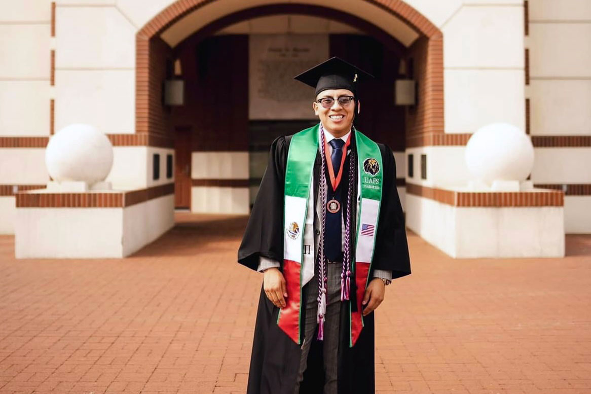 Student in cap and gown stands before the UAFS belltower