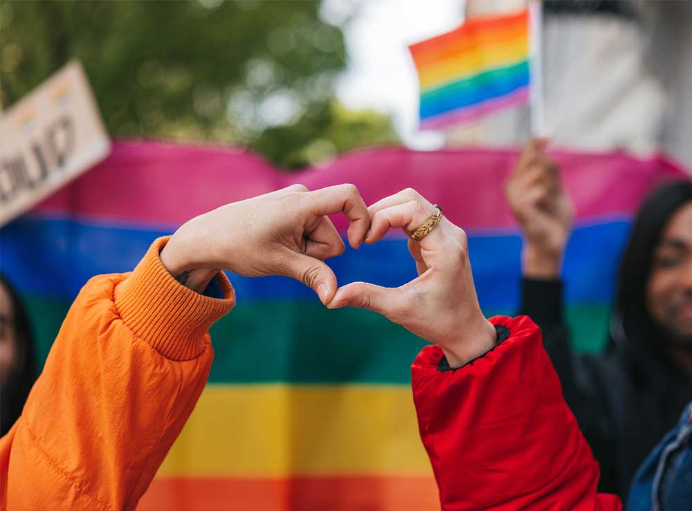 Hands form the shape of a heart in front of a rainbow flag