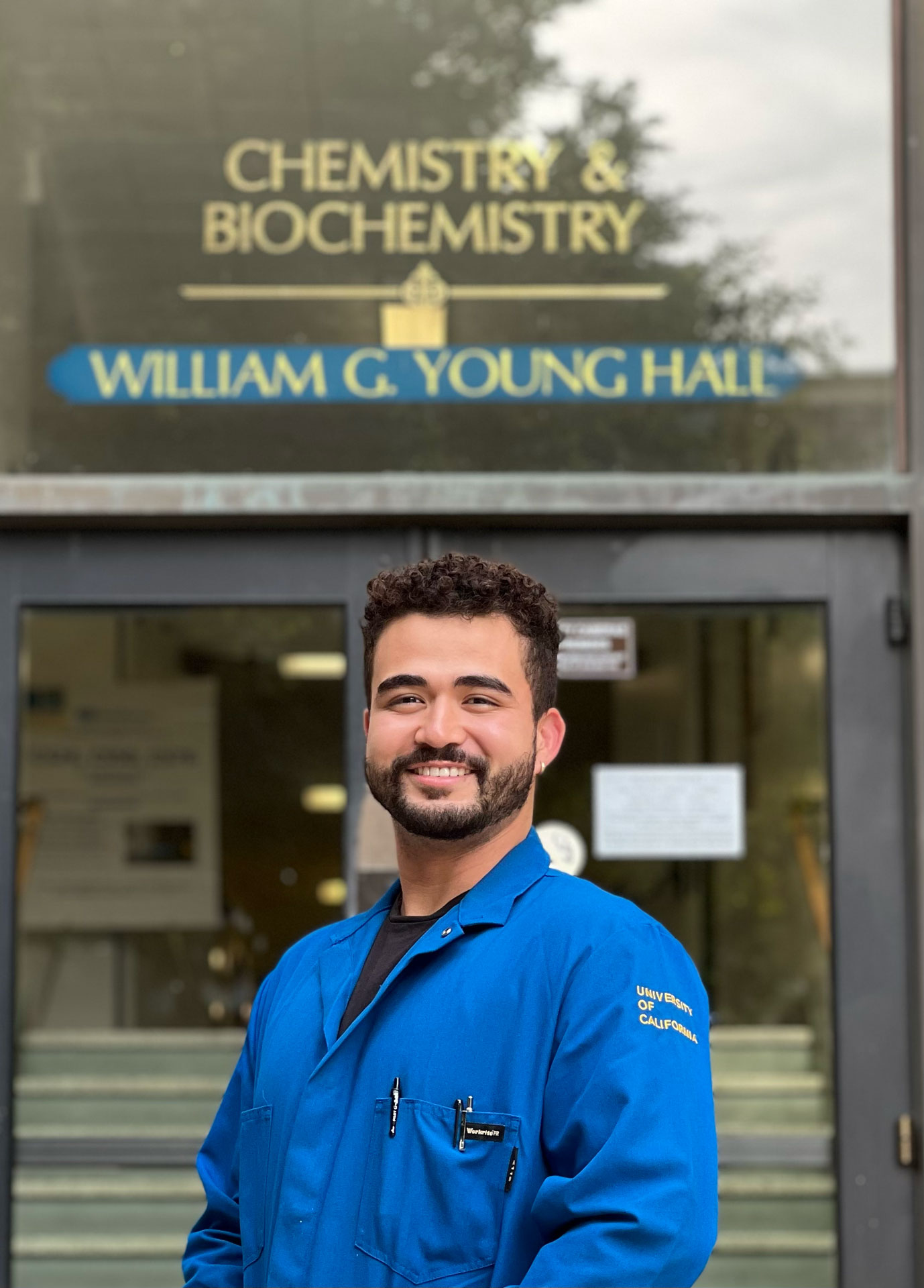 Individual in blue lab coat stands in front of glass entry to building. 