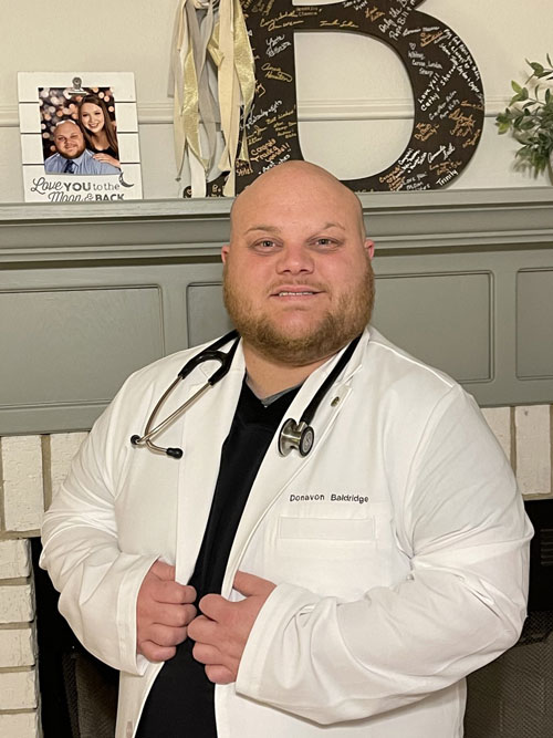 Individual in white lab coat smiles as he stands in front of a home fireplace. 