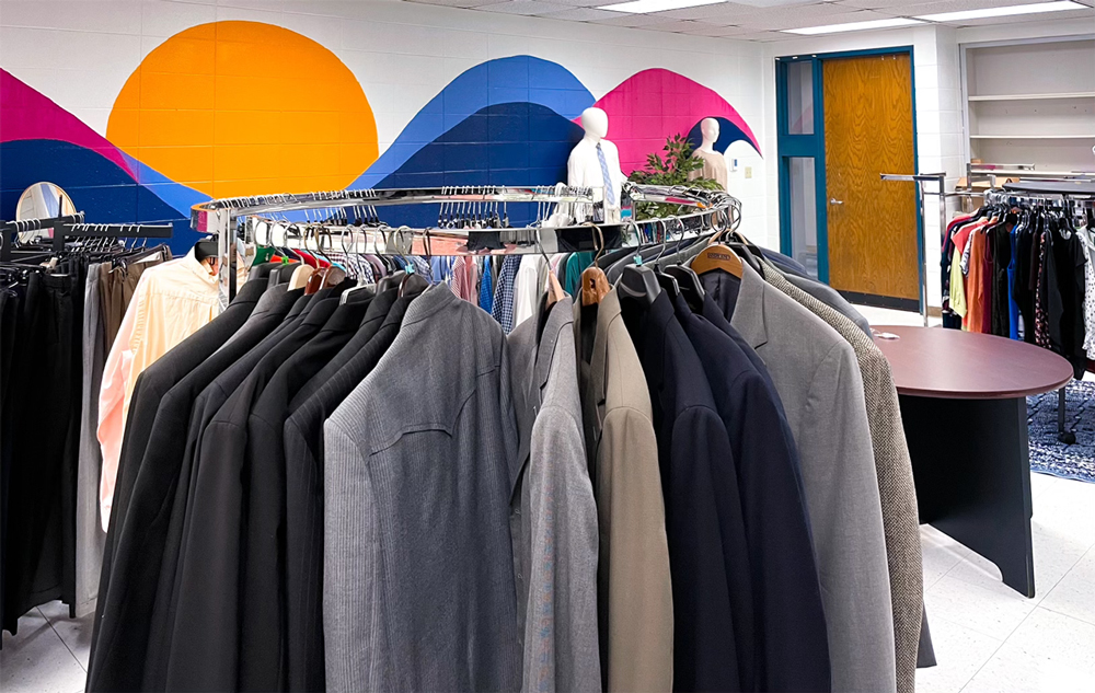 A mural of rolling hills and a sunrise is seen in the background with professional clothes on clothing racks in the foreground at the Babb Center for Career Services' Career Clothing Closet at UAFS.