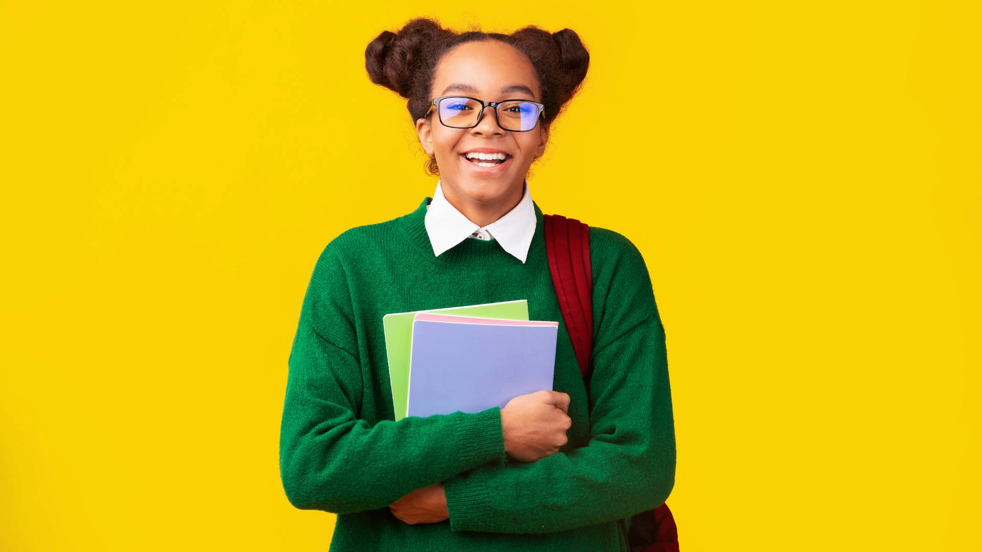 A girl holds a stack of books and a backpack in front of a yellow backdrop